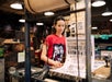 A woman in a red shirt operates a printing press, examining freshly printed posters in a workshop setting.