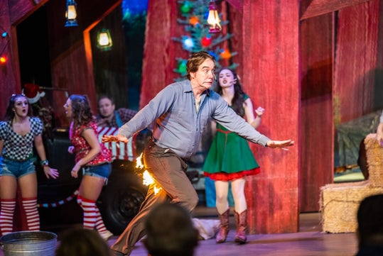 Man performing comically on stage with his pants on fire, surrounded by women in festive costumes, in a colorful theater set.