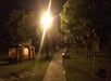 A paved path runs through a park at night, illuminated by a bright streetlamp, with benches, trees, and historical markers visible.