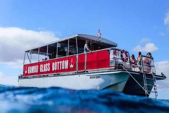 A glass-bottom boat labeled "Hawaii Glass Bottom" is on the water with several people on board, under a clear blue sky.