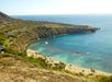 Aerial view of a crescent-shaped bay with clear blue water, sandy beach, and surrounding green hills under a clear sky.