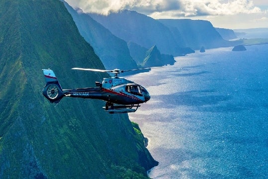 A silver and black Maverick airbus flying with the green rocky mountains and bright blue waters of the Island of Maui behind it.