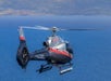 A gray and black helicopter with red accents flies over a large body of water, with land visible in the background.
