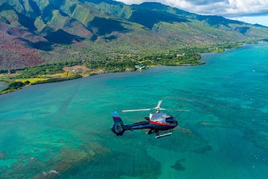 A helicopter flies over clear turquoise water near a lush, mountainous coastline under a partly cloudy sky.