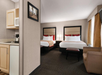 Hotel room with two beds, armchair, and side table. Room includes a kitchenette with microwave, fridge, and coffee maker. Neutral decor with red accents and framed wall art.