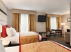 Hotel room with two double beds, a wall-mounted TV, a table with chairs, and a seating area. The room features neutral colors and decorative pillows.