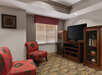 A cozy room with two red floral-pattern chairs, a wooden TV stand with a black TV, and a bookshelf. Carpeted floor and a window with closed blinds and a red plaid valance.