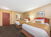 Hotel room with two double beds, white and gold bedding, a wall mirror, artwork, and a nightstand with lamps. The room has beige walls and a carpeted floor.
