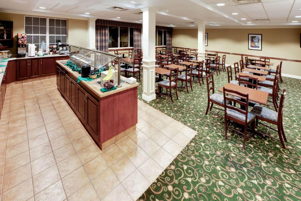 An empty dining area with green carpet and wooden tables and chairs. A buffet counter is on the left with various food items displayed. Large windows are present in the background.