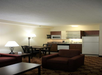 Cozy hotel room with a kitchenette, featuring a fridge, microwave, sink, and cabinets. There is a table with chairs, an armchair, and a lamp, all set on a patterned carpet.