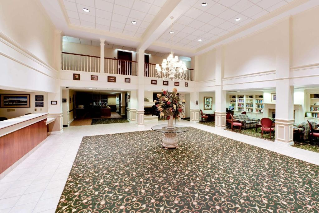 Spacious lobby with a floral arrangement in the center, surrounded by chairs and tables, featuring a chandelier above and a balcony with railings on the upper level.