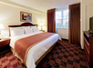 Hotel room with a king-sized bed, white and brown bedding, two bedside lamps, a window with curtains, framed art above the bed, and a TV on a wooden dresser.