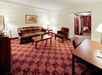 Hotel room with a patterned carpet, a plaid sofa, armchair, and coffee table. A desk with a chair is on the right. An open door reveals a bedroom with a bed and curtains.