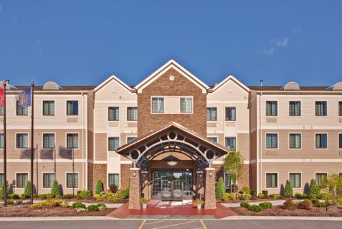 Front view of a three-story hotel building with a brick and beige facade, featuring a central entrance with an arched canopy. Flags are displayed to the left.