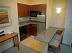 Compact kitchen with wooden cabinets, a small fridge, microwave, and dishwasher. A granite counter extends to a bar with two chairs. A painting hangs on the wall.