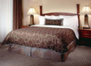 A neatly made bed with a brown patterned bedspread, white pillows, a decorative cushion, wooden headboard, and two bedside lamps on wooden nightstands in a carpeted room.
