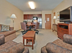 Hotel room lounge area with a dining table, chairs, sofa, armchairs, coffee table, flat-screen TV, and a kitchenette in the background.