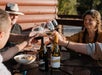 A group of friends smiling and cheering their wine glasses together at an outdoor table with Stomp By Croft Vineyards.