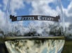 Close up of a wine glass with white wine in it and the logo for the Metzel Estate with a blue cloudy sky in the background.