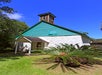 A small, white stone building with a green upper section and steeple, surrounded by grass and tropical plants, under a clear blue sky.