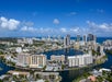 Aerial view of a coastal city with high-rise buildings, waterways, and the ocean in the background under a partly cloudy sky.