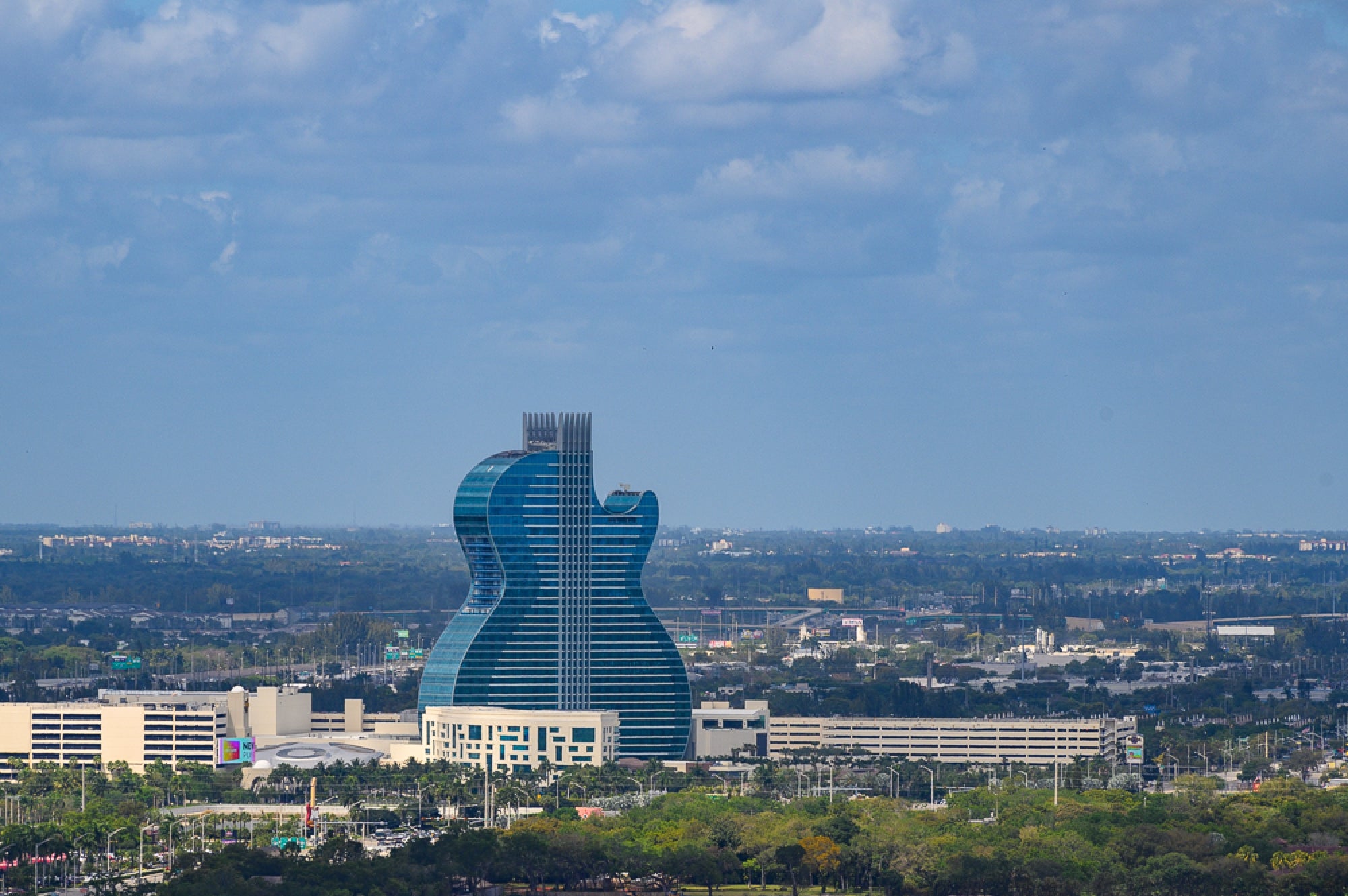 A large hotel building shaped like a blue guitar stands amid a cityscape under a partly cloudy sky.