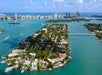 Aerial view of a narrow, residential island with houses and boats, surrounded by blue water, with city skyscrapers visible in the distance under a partly cloudy sky.