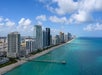 Aerial view of a city skyline with high-rise buildings along a sandy beach and turquoise ocean under a partly cloudy sky.