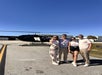 Four people stand together smiling on a tarmac in front of a black helicopter, with clear blue sky and hangars in the background.