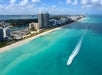 Aerial view of a city coastline with high-rise buildings, a sandy beach, turquoise water, and a motorboat creating a white wake offshore.