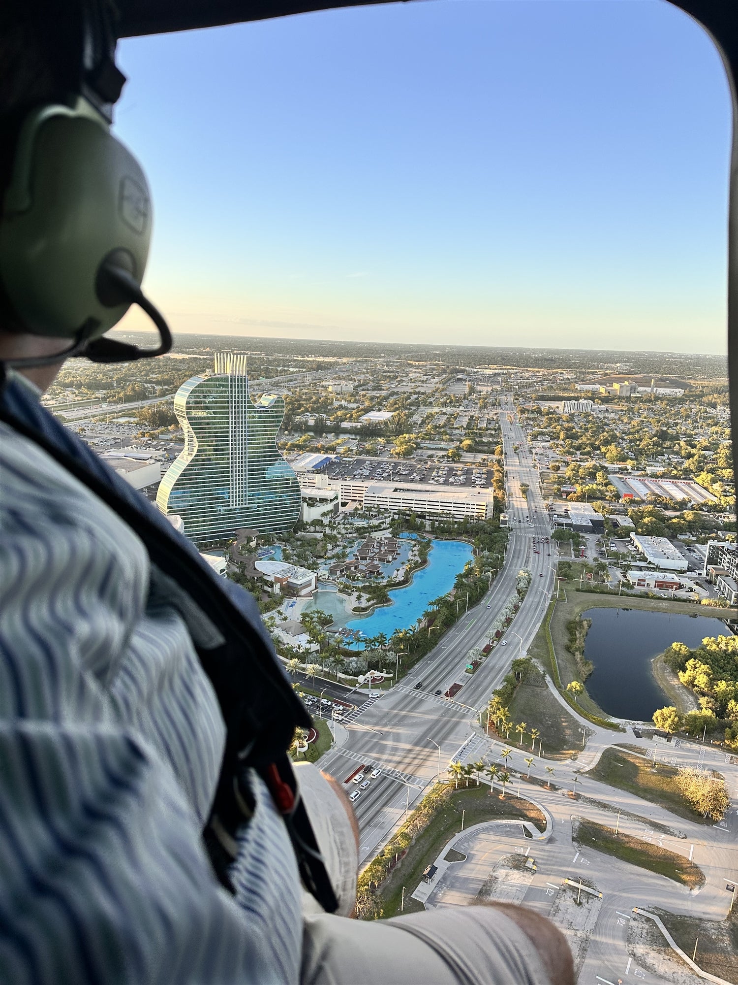 Person in a helicopter views a cityscape with a guitar-shaped building, roads, and pools below during daytime.