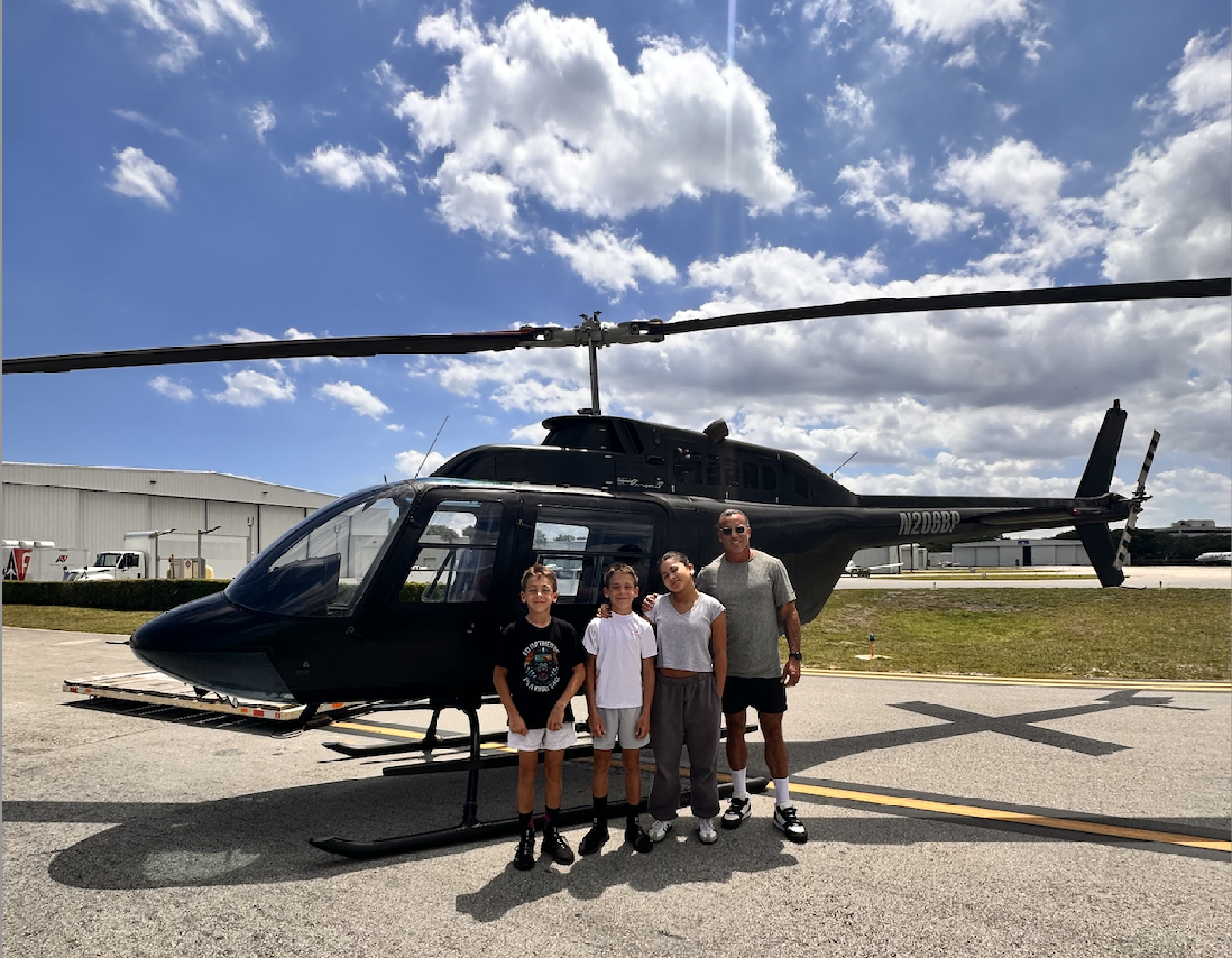Four people stand in front of a black helicopter parked on an airstrip, with a building and blue sky with clouds in the background.