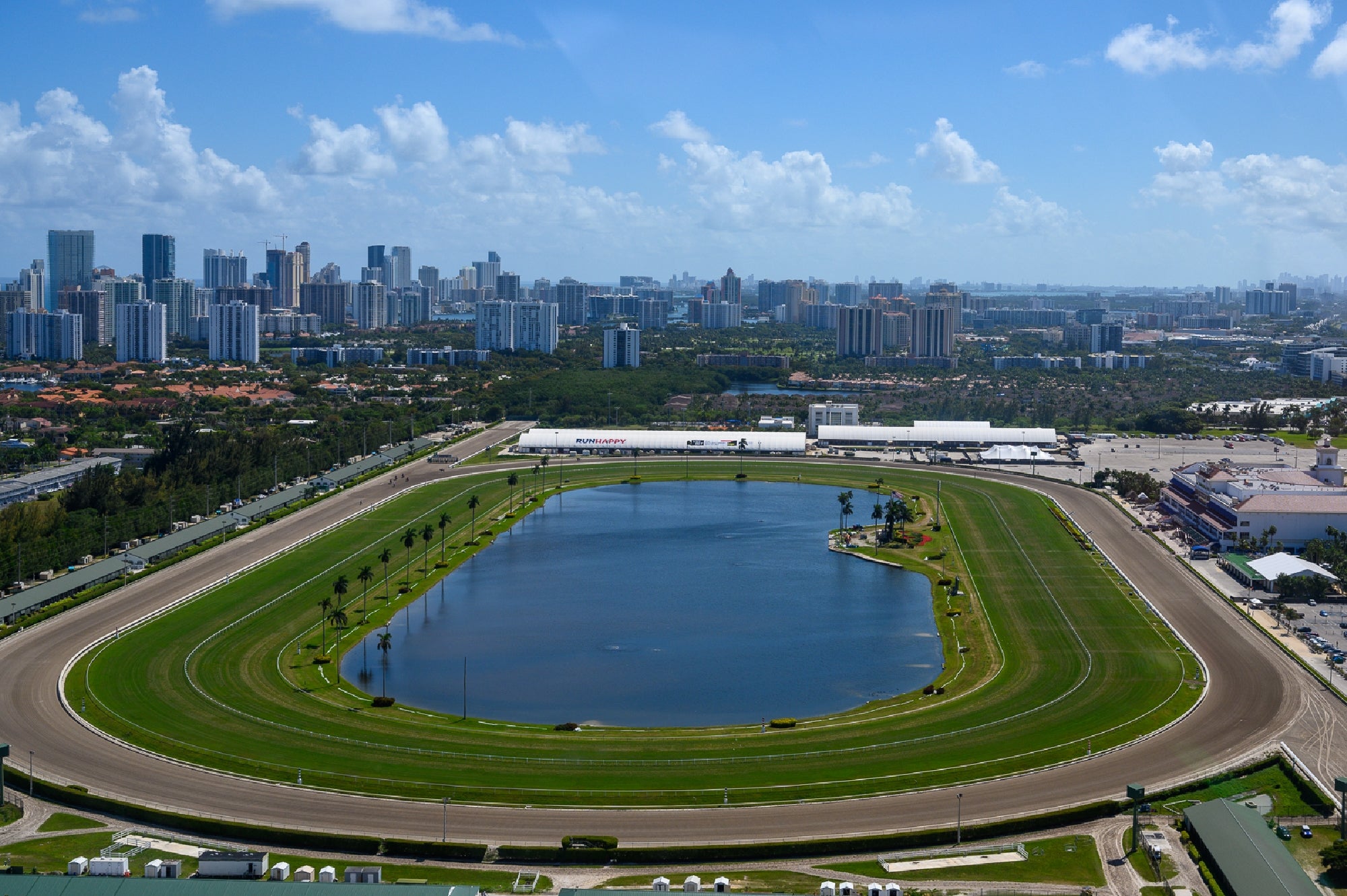 Aerial view of a large horse racing track with a lake in the center, surrounded by green grass, with city buildings in the background under a partly cloudy sky.