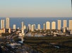 Aerial view of a coastal city with high-rise buildings, waterways, and dense greenery in the foreground under clear skies.