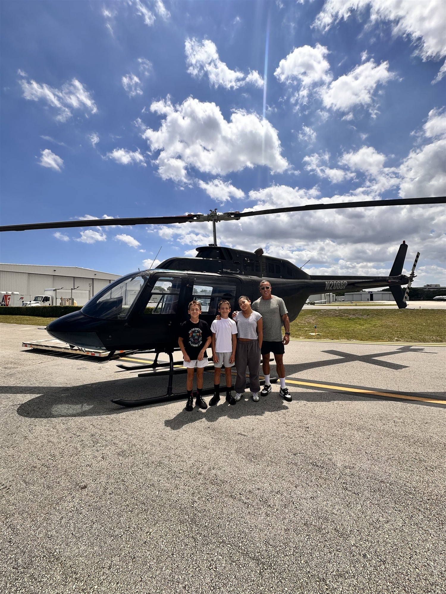 Four people stand together in front of a black helicopter on an airport tarmac under a partly cloudy sky.