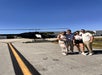 Four people stand together on an airport tarmac in front of a black helicopter on a sunny day.