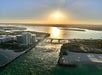 Aerial view of Haulover beach at sunset, showing high-rise buildings, a bridge over water, and a sandy beach with the sun reflecting on the sea.