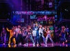 A vibrant stage performance with a group of dancers in colorful outfits, under dynamic lighting and an urban backdrop.