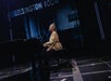 A person seated at a piano on a dark stage, with "Ellington Room" displayed above them.