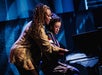Two musicians at a piano; one plays while the other gestures, likely instructing. They are on a stage with a blue-lit backdrop.