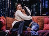 Two people sitting on a red sofa, embracing and smiling warmly. The background features a cityscape at night.
