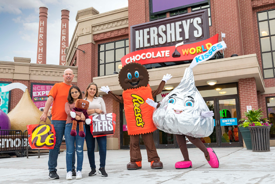 A family poses with Reese's and Hershey's Kiss mascots in front of a Hershey's Chocolate World entrance.