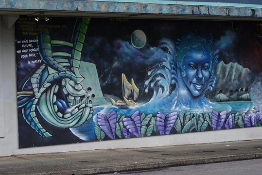 A mural depicts a blue robotic figure, a sailing canoe, a large blue human face among waves, purple taro leaves, mountains, and a moonlit sky with a quote attributed to B. Marley.