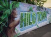 A mural on corrugated metal features the words "Hilo Town" in large green letters next to a painted profile of a person and green leaves.