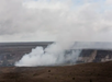 A volcanic crater emits a plume of white smoke under a cloudy sky, surrounded by barren landscape.