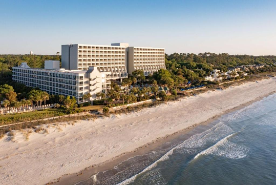 Aerial view of a large beachfront hotel surrounded by trees, with the ocean and sandy beach in the foreground under a clear blue sky.