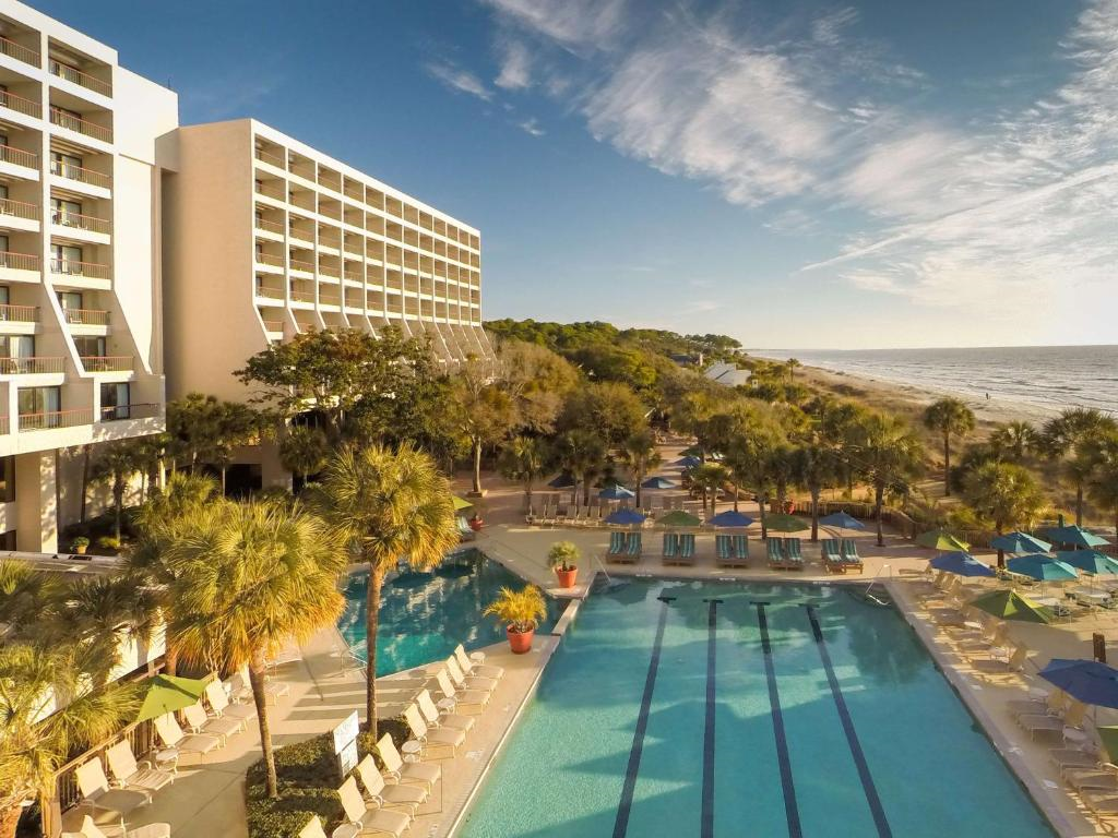 Aerial view of a beachfront hotel with swimming pools, sun loungers, and palm trees, situated next to the ocean under a partly cloudy sky.