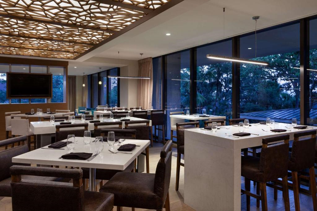 Modern restaurant interior with white tables set with glassware and dark napkins. Large windows provide a view of greenery. Ceiling features decorative geometric design.