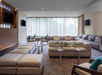 A modern lounge area with beige seating, small round tables, TVs on the walls, and large windows with sheer curtains.