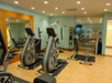 A gym room with exercise machines, free weights, a large mirror, and a stability ball. The room has wooden flooring and light-colored walls.